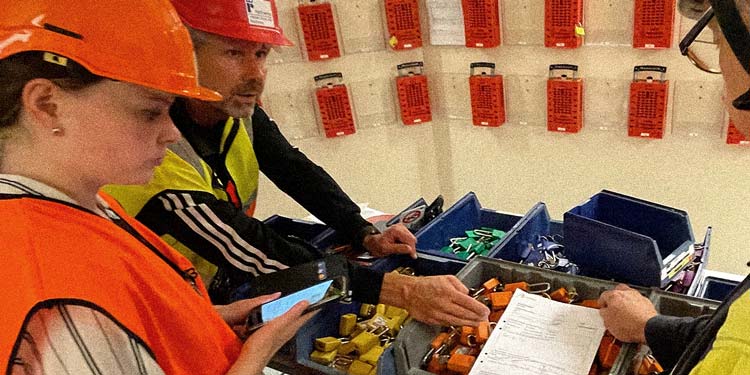 Personer i skyddsväst och hjälm hanterar pappersformulär och mobiltelefon bland lådor med teknisk utrustning